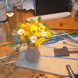 Un bouquet de fleurs jaunes et blanches mixtes sur un table de travail avec des baguettes de verre de Murano et des outils à façonner le verre aux alentours. 